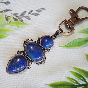 May include: A copper key chain with three lapis lazuli stones. The stones are oval and teardrop shaped and are set in a decorative copper frame. The key chain has a lobster claw clasp.