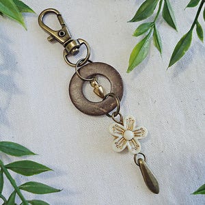 May include: A brown wooden circle with a gold metal ring and a small gold metal charm attached to a white flower charm with gold accents. The charm is attached to a gold metal key chain.