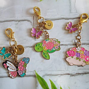 May include: Three gold-colored keychains with decorative charms. One features a butterfly with pink, white, and teal accents, another a green turtle with floral details, and the last a pink hedgehog with flowers. Each charm has a gold initial.