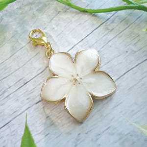 May include: A white flower charm with a gold-colored border and clasp. The flower has five petals and a textured center. The charm is likely made of metal and enamel, and is a decorative accessory.