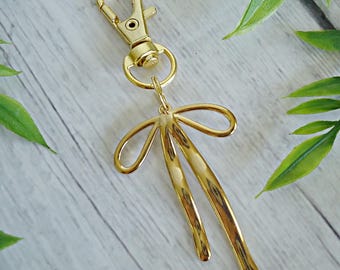 May include: A gold-colored keychain with a lobster clasp and a decorative bow-shaped charm. The charm has a stylized design with curved lines and a polished finish. The keychain is shown against a light wood-grain background with green leaves.