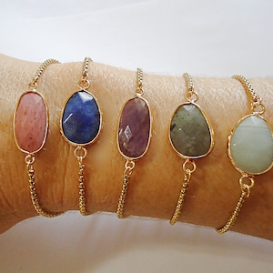 May include: Five gold chain bracelets with different colored oval-shaped gemstones. The bracelets are on a person's wrist. The gemstones are pink, blue, purple, green, and light green.