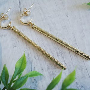 May include: A pair of gold-colored earrings with a textured, linear design. Each earring features a small hoop that connects to a long, textured bar. The earrings are displayed on a light-colored surface with green foliage.