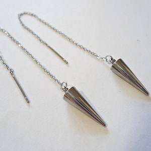 May include: A pair of silver-toned, cone-shaped dangle earrings. Each earring features a long chain that threads through the ear, with a small bar at one end and a cone pendant at the other. The earrings are displayed on a white background.