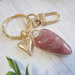 May include: A gold-colored keychain with a heart-shaped charm and a larger, teardrop-shaped charm with a pinkish-red stone. The keychain includes a clasp and a small heart charm.