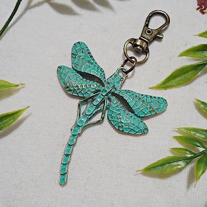 May include: A turquoise blue dragonfly keychain with a bronze-colored metal clasp. The dragonfly has a detailed, textured design.