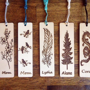 May include: Five wooden bookmarks with intricate pyrography designs. Each bookmark features a different image: a floral branch, birds on a branch, a feather, a pine tree, and a dragon. Each bookmark has a tassel and a name: Mom, Lydia, Alana, and Cora.