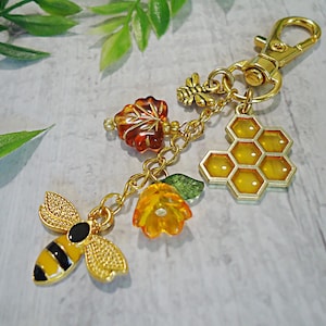 May include: Gold-colored keychain with bee, honeycomb, and flower charms. The bee charm has black and yellow stripes. The honeycomb charm is yellow. The keychain also features a leaf and flower charm. The clasp is gold.