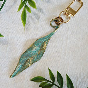 May include: A leaf-shaped keychain with a teal and gold finish. The metal leaf has detailed veining and is attached to a gold-colored clasp and key ring. The background is a white fabric with green leaves.