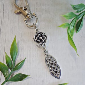 May include: Silver-toned keychain with a lobster clasp, a floral bead, and a Celtic knot pendant. The keychain is made of metal and features intricate details. Green leaves are in the background.