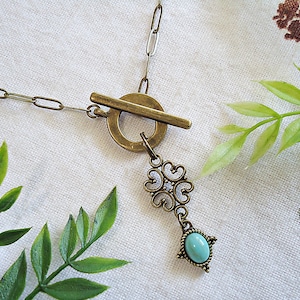 May include: A vintage-style necklace with a bronze-colored chain and clasp. The pendant features an ornate design with a turquoise-colored stone at the bottom. The necklace is displayed on a white fabric background with green leaves.