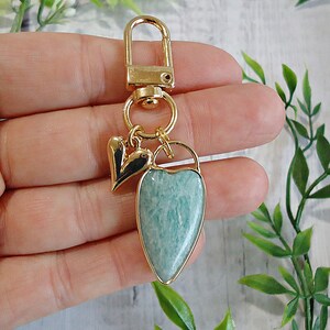 May include: A gold-colored keychain with a heart-shaped pendant made of a light blue stone. The pendant is framed in gold and has a gold-colored clasp. A small black and gold heart charm hangs next to the pendant.