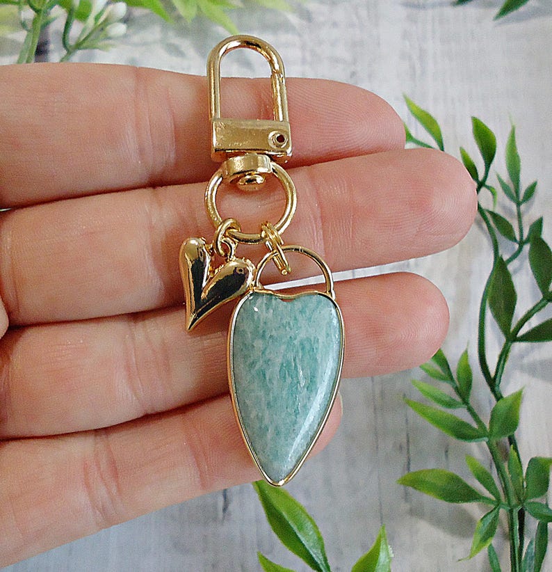 May include: A gold-colored keychain with a heart-shaped pendant made of a light blue stone. The pendant is framed in gold and has a gold-colored clasp. A small black and gold heart charm hangs next to the pendant.