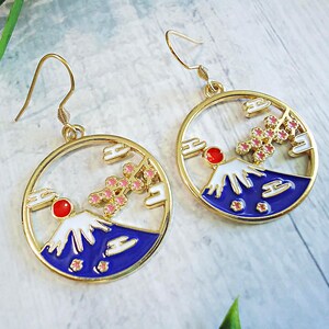 May include: A pair of gold-toned circular drop earrings featuring an enamel design of a mountain with a white snow-capped peak, a deep blue base, a bright red sun, and pink cherry blossom branches, suspended from simple gold-toned metal hooks.