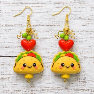 May include: A pair of dangle earrings featuring a yellow taco charm with a smiling face, topped with a bright red heart bead and a small green bead. The charms are suspended from gold-toned metal hooks with coiled wire accents and small loops.