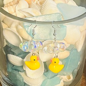 May include: A pair of yellow rubber ducky earrings with iridescent bubble charms hanging from silver hooks. The earrings are displayed in a glass jar filled with seashells and blue glass.