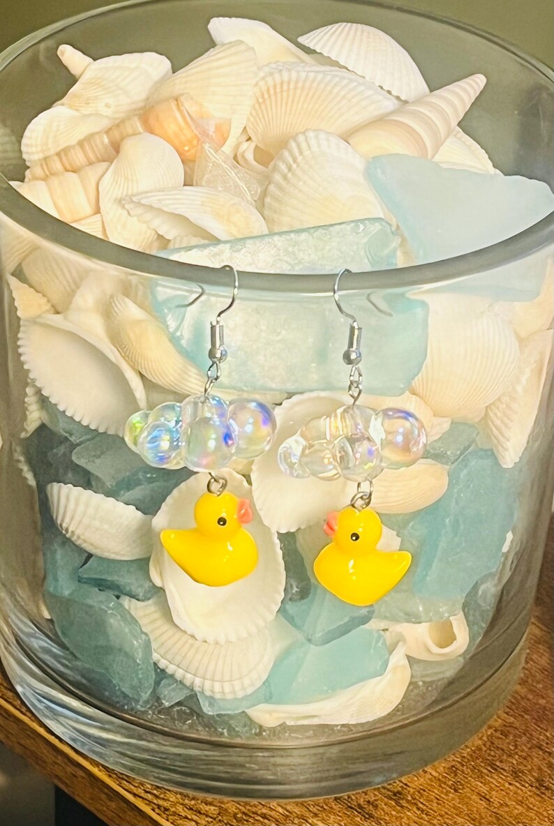 May include: A pair of yellow rubber ducky earrings with iridescent bubble charms hanging from silver hooks. The earrings are displayed in a glass jar filled with seashells and blue glass.