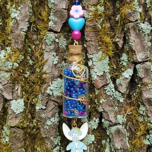 May include: A whimsical necklace featuring a small glass bottle filled with blue and pink beads, adorned with gold wire and a fairy charm. The necklace has a black cord and is accented with pink and blue beads.