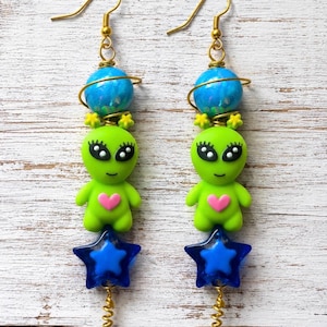 May include: A pair of dangle earrings featuring a gold-tone hook, a blue planet bead with a gold ring, a lime green alien figure with a pink heart on its chest, and a dark blue star-shaped bead at the bottom with a small gold spiral wire tail.