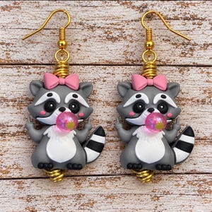 May include: A pair of dangle earrings featuring gray raccoon charms with white bellies, black and white striped tails, and pink bows on their heads. Each raccoon is blowing a pink bubble and holding up its middle fingers. Gold-tone metal hooks.