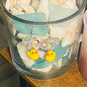 May include: A pair of yellow rubber ducky earrings with iridescent beads. The earrings are hanging from silver hooks.