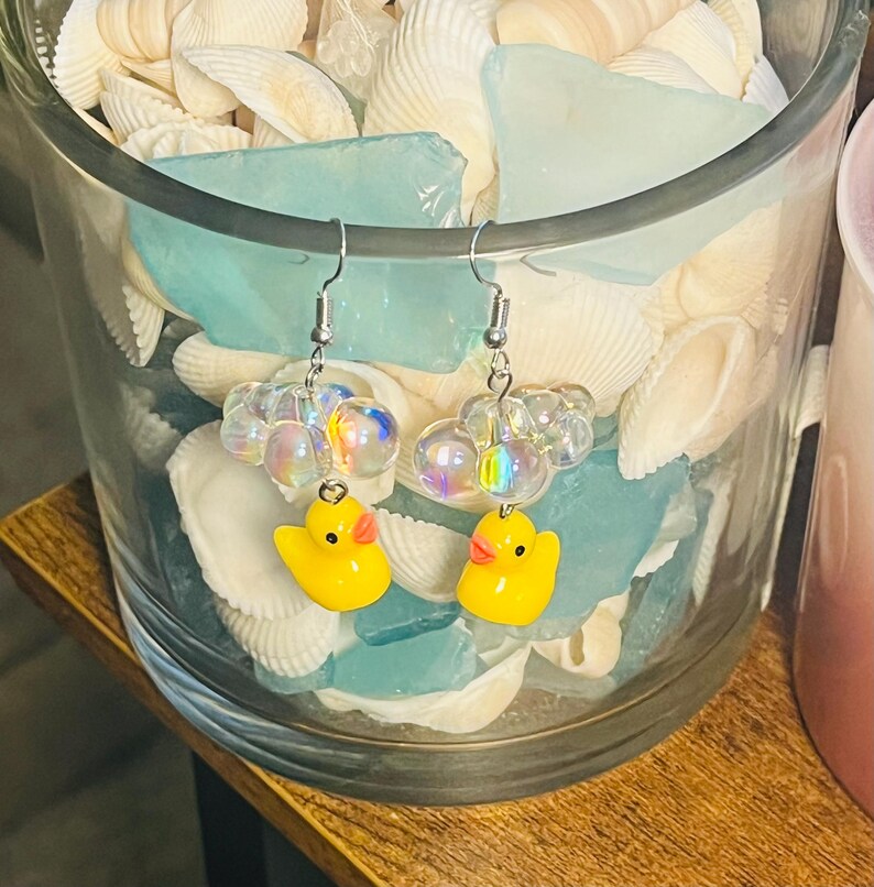 May include: A pair of yellow rubber ducky earrings with iridescent beads. The earrings are hanging from silver hooks.