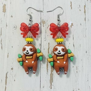May include: A pair of handmade earrings featuring a brown sloth charm holding a branch with green leaves. Each earring has a red bow and yellow bead above the sloth, with a silver fish hook.