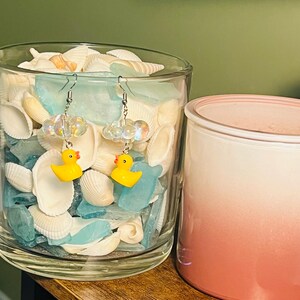 May include: A pair of yellow rubber ducky earrings with clear iridescent beads and silver hooks. The earrings are displayed in a glass jar filled with seashells.