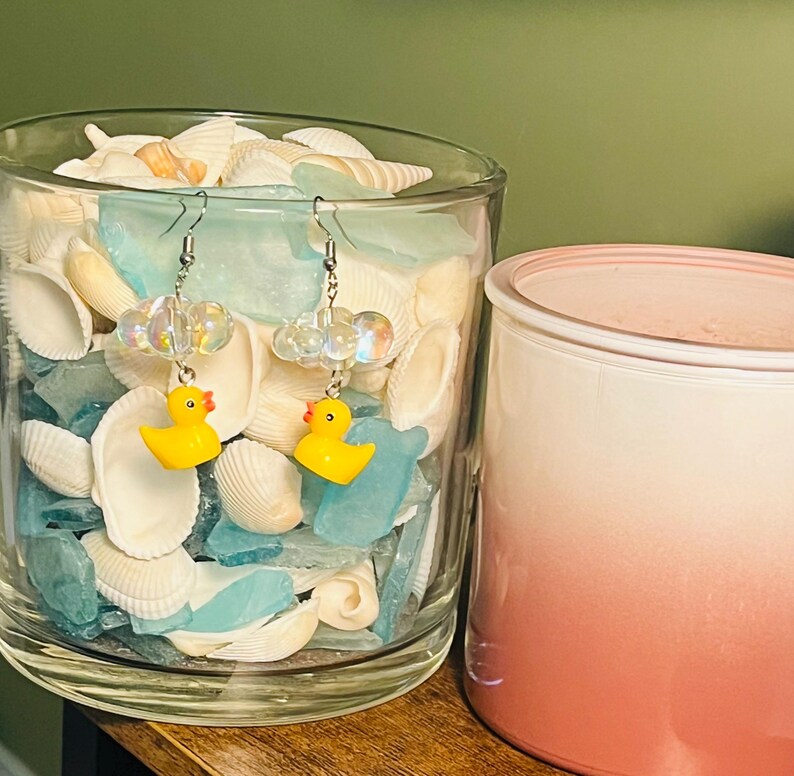 May include: A pair of yellow rubber ducky earrings with clear iridescent beads and silver hooks. The earrings are displayed in a glass jar filled with seashells.
