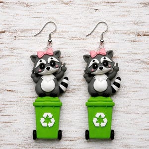 May include: A pair of dangle earrings featuring a gray and white raccoon with a pink bow on its head, standing on a green recycling bin. The raccoon is depicted with its middle finger raised on each hand. The earrings have silver-toned hooks.