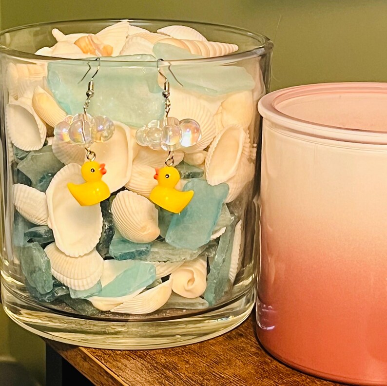 May include: A pair of yellow rubber ducky earrings with clear iridescent beads and silver hooks. The earrings are displayed in a glass jar filled with seashells and blue glass.