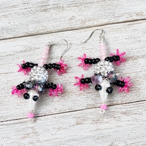 May include: A pair of dangle earrings shaped like lizards, crafted from black, white, and pink beads. The lizard bodies feature sparkling silver-toned beads, black bead limbs, and pink star-shaped accents, suspended from silver-toned metal hooks.