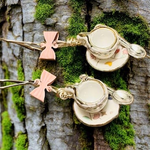 May include: Two miniature tea cup and saucer charms with gold-colored spoons and a copper-colored bow. The teacups are white with a gold rim and sit on a small saucer with floral designs. The charms are attached to a gold-colored wire.