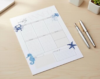 May include: A weekly planner with a watercolour design featuring a crab, jellyfish, seahorse, and starfish in shades of blue. Two silver pens are placed next to the planner on a light wood surface.