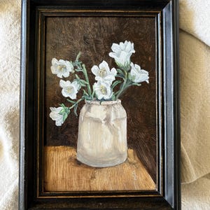 May include: An oil painting in a black frame depicting a white ceramic jar filled with white bell-shaped flowers. The background is a dark brown textured surface, and the jar sits on a light brown wooden table. The artist's initials are visible.