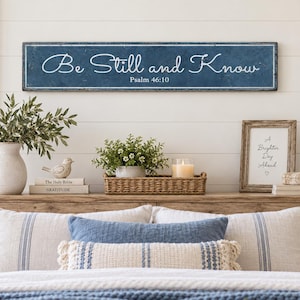 May include: A rectangular blue wooden wall sign with white distressed edges and the white cursive text Be Still and Know Psalm 46:10. The sign is mounted above a bed featuring white and blue pillows, a blue knit throw, and a wooden headboard.