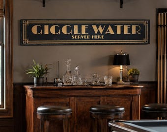 Op de afbeelding: Een rechthoekig zwart wandbord met gouden letters met de tekst GIGGLE WATER SERVED HERE, voorzien van een geometrisch randontwerp. Het bord is aan de muur bevestigd boven een houten barkast met twee krukken met lederen zitting en glaswerk.