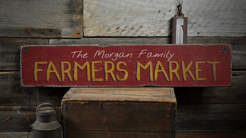 Puede incluir: Un letrero de madera rojo con texto blanco que dice "The Morgan Family Farmers Market" en una fuente r&uacute;stica. El letrero est&aacute; desgastado y tiene un aspecto desgastado.