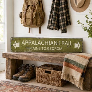 May include: A rectangular olive green wooden sign with distressed edges featuring the white text APPALACHIAN TRAIL and MAINE TO GEORGIA. The sign includes white directional arrows on both ends, each marked with the letters S and N respectively.