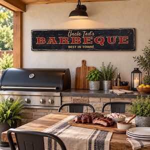 May include: A rectangular black wooden sign with distressed edges featuring the red and white text Uncle Ted's BARBEQUE BEST IN TOWN! mounted on a light-colored wall above a stainless steel outdoor grill and a wooden dining table with food.