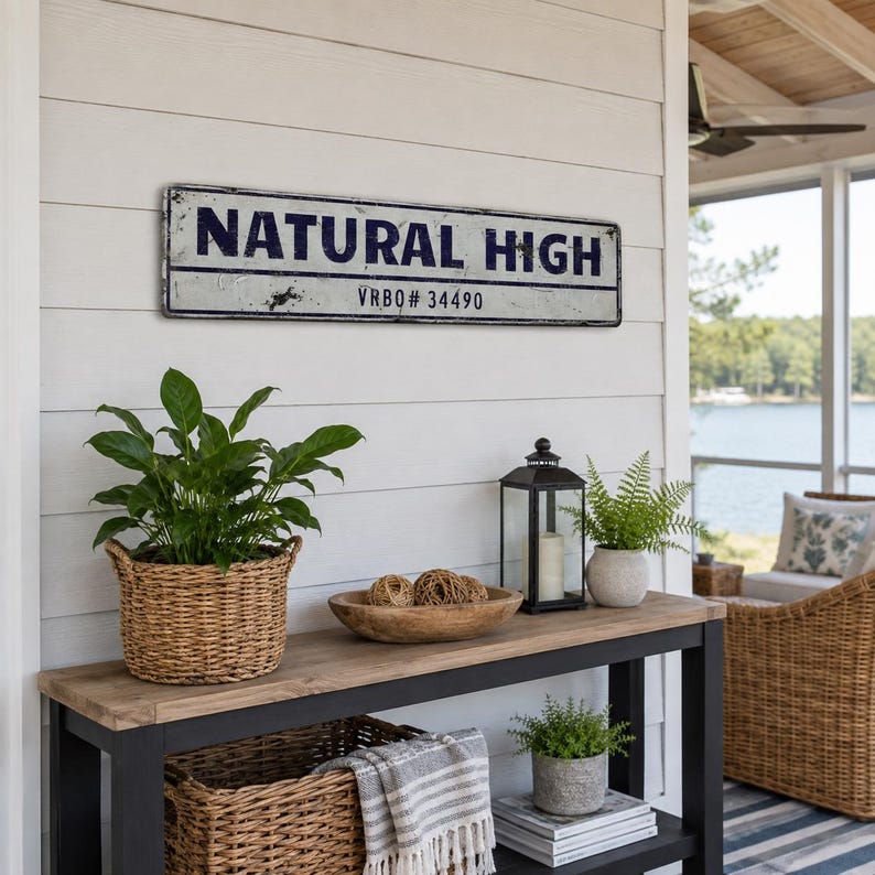 May include: A rectangular metal sign with a white background and dark blue text reading NATURAL HIGH and VRBO# 34490. The sign features a distressed finish with visible wear along the edges and surface, mounted on a white horizontal paneled wall.