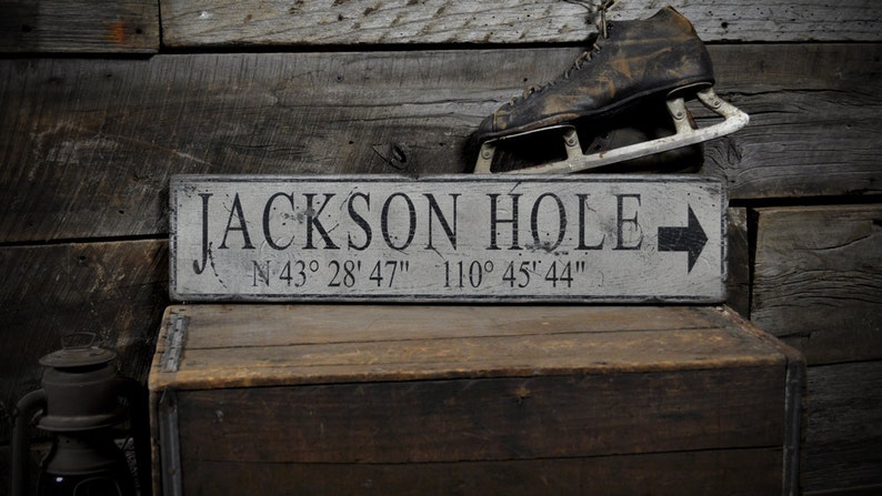 May include: A rectangular wooden sign with a distressed white finish and black lettering that reads JACKSON HOLE followed by the coordinates N 43&deg; 28' 47" 110&deg; 45' 44" and a black arrow pointing to the right side of the sign.