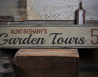 Puede incluir: Un letrero de madera desgastado con pintura blanca y letras negras dice "Aunt Bethany's Garden Tours 5¢". El letrero está colgado en una pared de madera rústica.