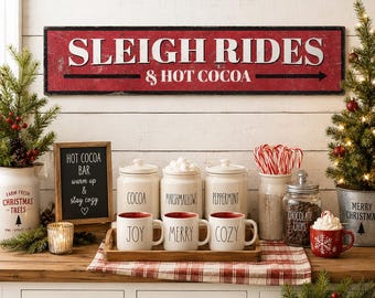 May include: A rectangular red sign with white text reading SLEIGH RIDES & HOT COCOA, featuring a black arrow. Below, three white ceramic mugs labeled JOY, MERRY, and COZY sit on a wooden tray alongside three matching canisters for cocoa.