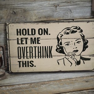 May include: A rustic wooden sign with a black and white image of a woman thinking. The sign reads "Hold on. Let me overthink this."