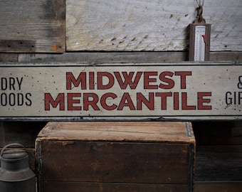 Op de afbeelding: Een vintage houten bord met witte verf en rode letters met de tekst "Midwest Mercantile Dry Goods & Gifts".