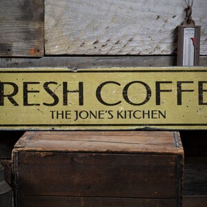May include: A yellow wooden sign with black text that reads "FRESH COFFEE THE JONE'S KITCHEN". The sign is hanging on a rustic wooden wall.