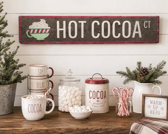 Pode incluir: Uma placa retangular de madeira com o texto HOT COCOA CT e um gráfico de uma caneca com chantilly. A exibição inclui canecas empilhadas, um pote de vidro com marshmallows, uma lata de metal para cacau, bengalas doces e um sinal WARM UP HERE.