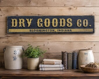 Op de afbeelding: Een rechthoekig houten bord met een donkerblauwe achtergrond en gele letters met de tekst DRY GOODS CO. BLOOMINGTON, INDIANA. Het bord heeft een verweerde afwerking en is bevestigd boven een houten oppervlak met twee keramische potten.