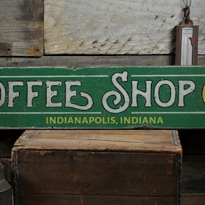 Może przedstawiać: Zielony drewniany znak z białym tekstem, który brzmi "Coffee Shop" i "Indianapolis, Indiana". Na prawej stronie znaku znajduje się żółte koło z czarnym tekstem, który brzmi "Now Open!"